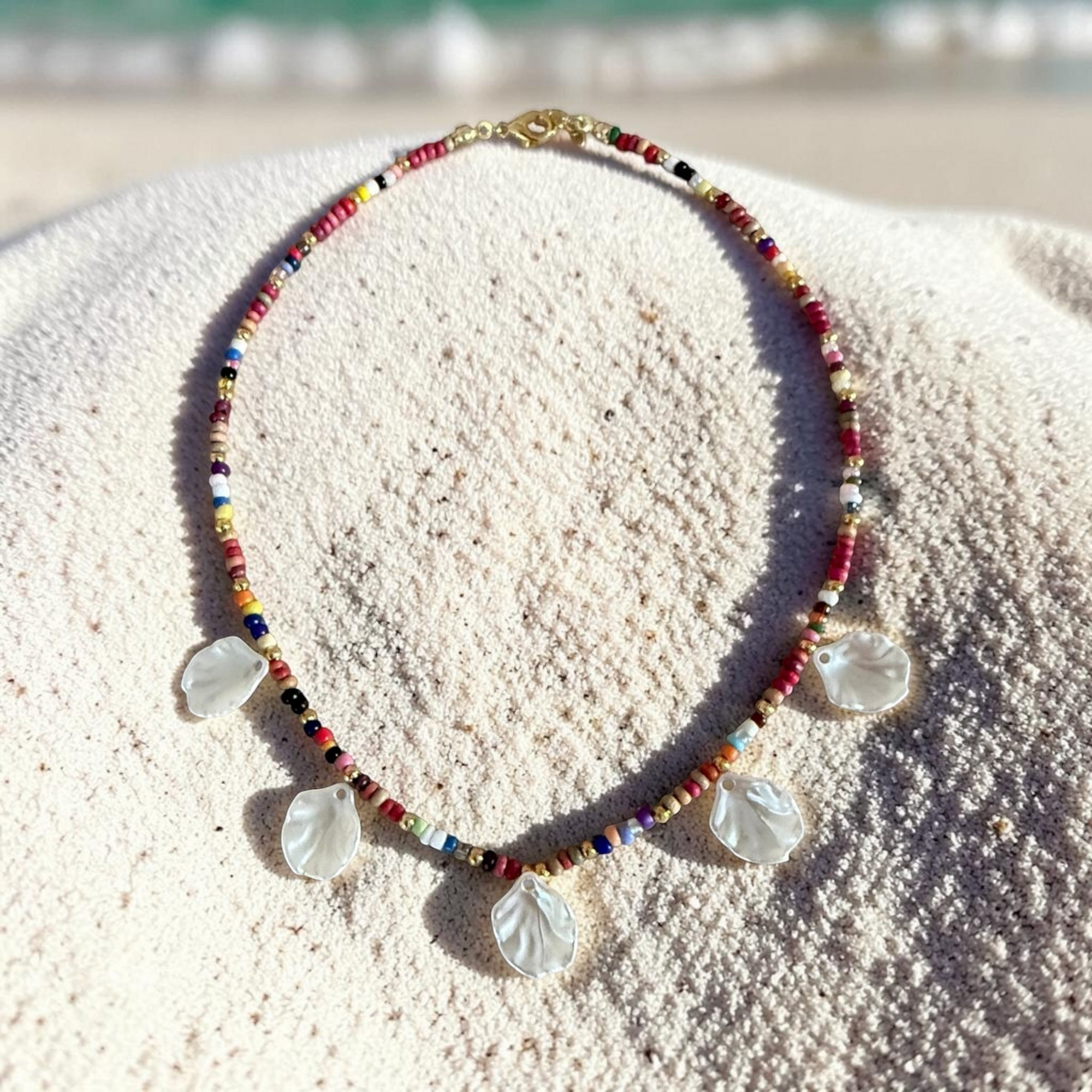 Craft and Babble beaded necklace with multi-coloured glass beads and five iridescent shell charms on sandy beach.