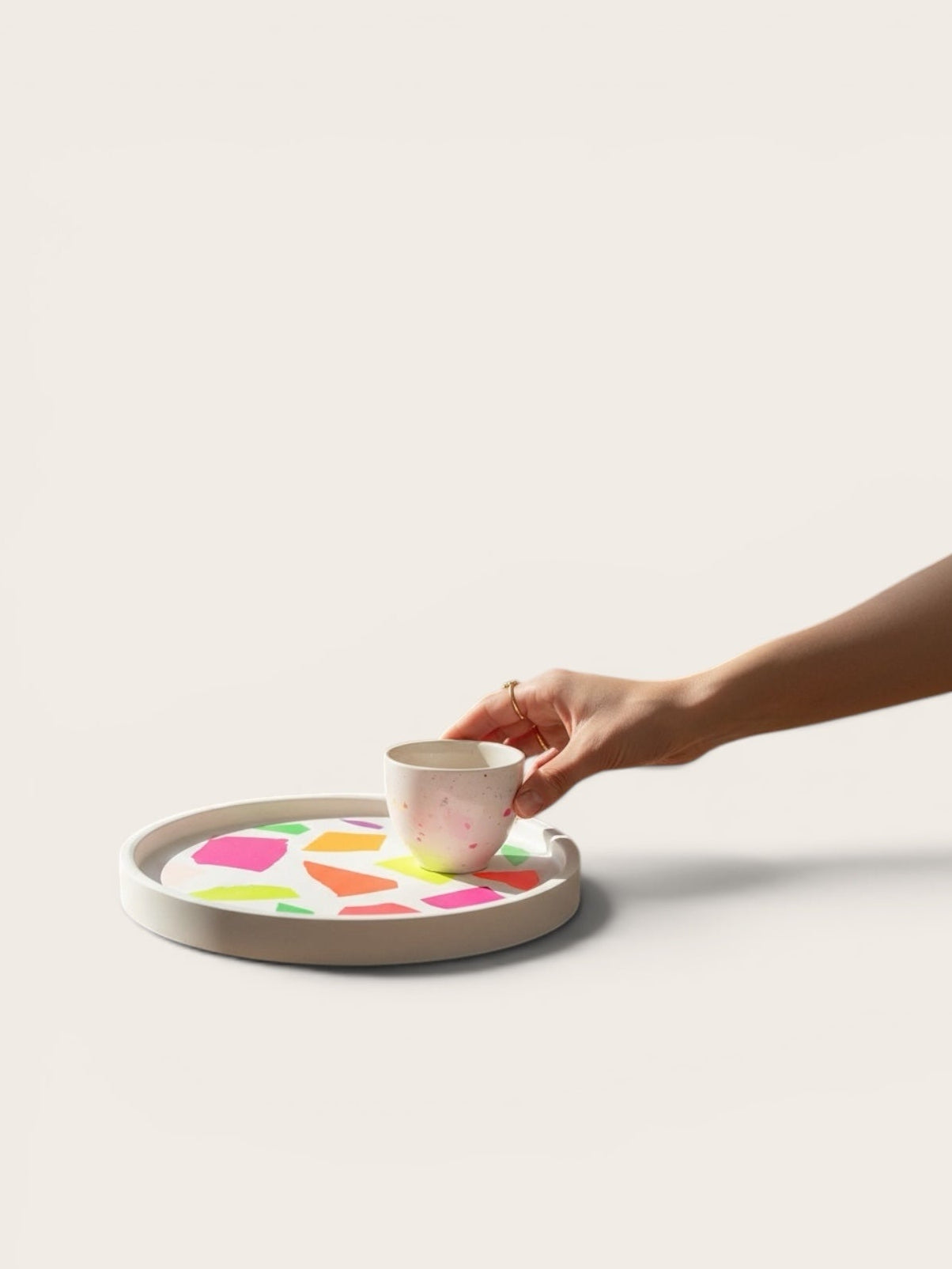 Craft and Babble circular white serving tray with vibrant neon terrazzo pattern, held by a hand with a small cup.